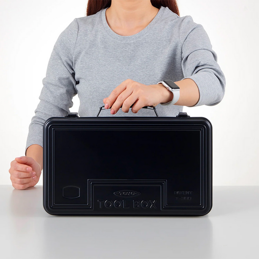 Toyo Steel T-360 Black. Glossy black steel toolbox with a round handle being held by a person. Designed for durability, stackable, and features a cylinder lock. 1.6kg.